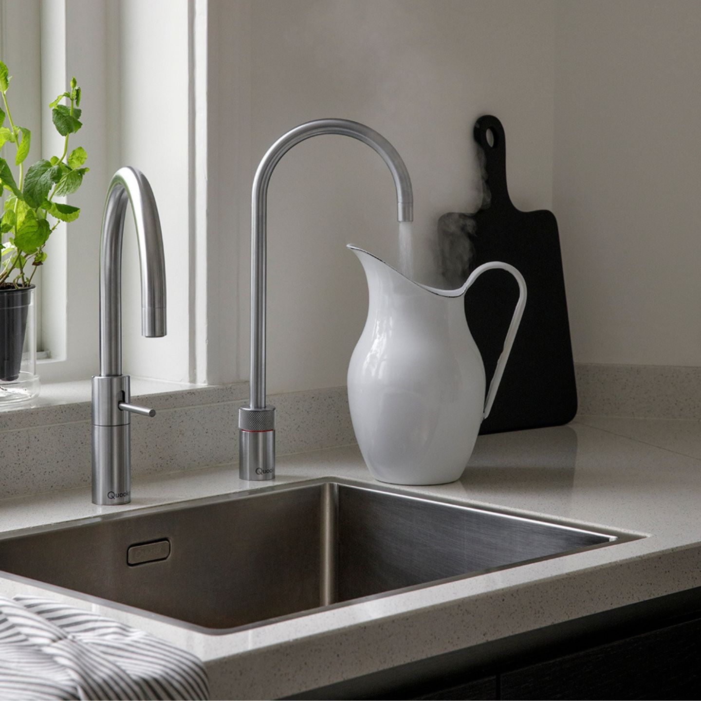 Ein dampfender weißer Keramikkrug steht unter einem modernen Wasserhahn auf einer Küchentheke neben einem Waschbecken, mit einer Topfpflanze und einem Schneidebrett im Hintergrund.