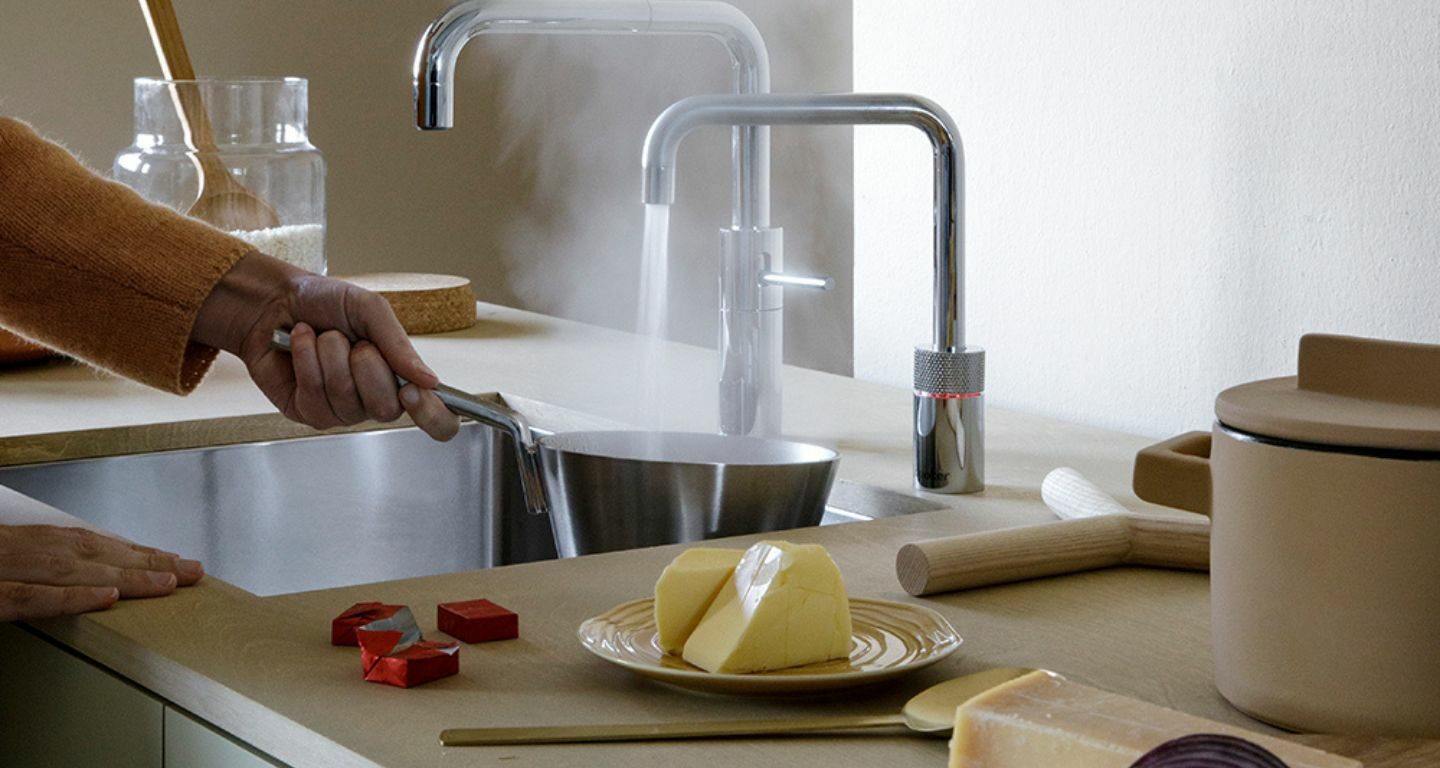Eine Person hält einen dampfenden Topf unter einen modernen Küchenhahn, auf der Arbeitsplatte liegen Käse, eine Zwiebel und Küchenutensilien.