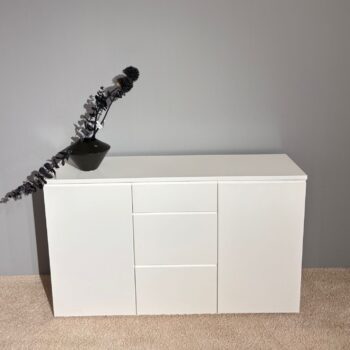 Das Sideboard Optima 3 mit drei Schubladen in der Mitte steht auf einem beigen Teppich vor einer grauen Wand; darauf steht eine schwarze Vase mit dunklem, angelehntem Laub.