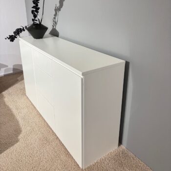 Das Sideboard Optima 3 zeichnet sich durch klare Linien aus und steht vor einer hellgrauen Wand auf einem beigen Teppich. Eine schwarze Vase mit Zweigen schmückt die obere linke Ecke und verleiht diesem stilvollen Sideboard einen modernen Touch.