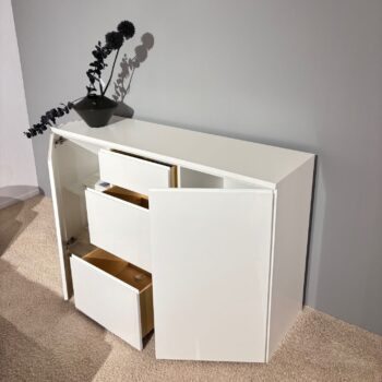 Die Anrichte Optima 3 mit drei Schubladen und einer Tür, die teilweise geöffnet ist, steht auf einem beigen Teppich vor einer grauen Wand und wird von einer schwarzen Vase mit dunklen Trockenblumen gekrönt.