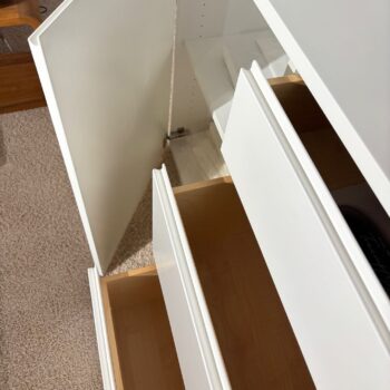 Das Sideboard Optima 3 besteht aus einer weißen Kommode mit zwei teilweise geöffneten Schubladen und einer angelehnten Tür, die den Blick auf das helle Holzinnere freigibt. Die Anrichte steht auf einem beigen Teppich neben einem Holzboden.