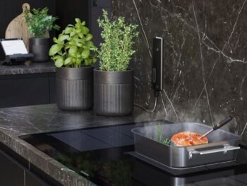 Siemens: Eine moderne Küche mit einer Arbeitsplatte aus schwarzem Marmor, getopften Kräutern, einem Schneidebrett und einer Pfanne mit Speisen, die auf einem Induktionsherd kochen.