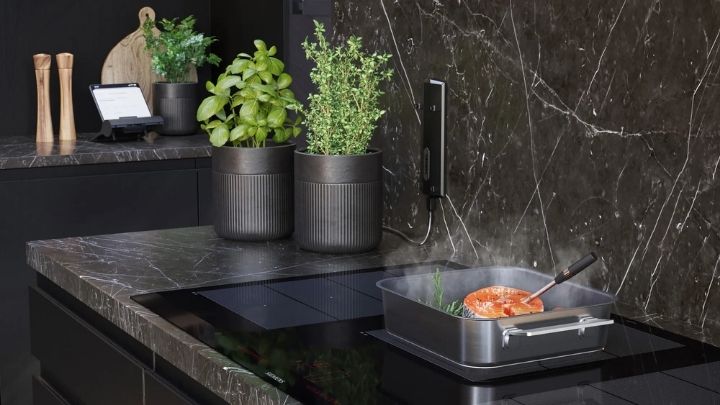 Siemens: Eine moderne Küche mit einer Arbeitsplatte aus schwarzem Marmor, getopften Kräutern, einem Schneidebrett und einer Pfanne mit Speisen, die auf einem Induktionsherd kochen.