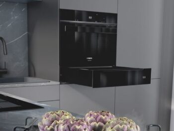 Siemens: Eine moderne Küche mit einem schwarzen Einbaubackofen mit einer ausziehbaren Schublade, einer grauen Arbeitsplatte und Artischocken, die in einem Topf auf dem Herd dampfen.