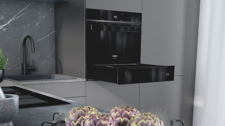 Siemens: Eine moderne Küche mit einem schwarzen Einbaubackofen mit einer ausziehbaren Schublade, einer grauen Arbeitsplatte und Artischocken, die in einem Topf auf dem Herd dampfen.