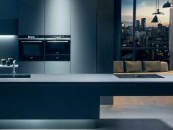 Siemens: Moderne Küche mit eleganten dunklen Schränken, eingebauten Öfen, einer Kaffeemaschine und einer minimalistischen Insel vor großen Fenstern, die die Skyline der Stadt in der Abenddämmerung zeigen.