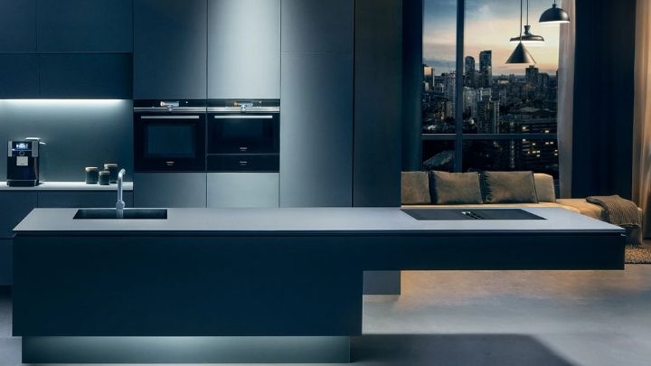 Siemens: Moderne Küche mit eleganten dunklen Schränken, eingebauten Öfen, einer Kaffeemaschine und einer minimalistischen Insel vor großen Fenstern, die die Skyline der Stadt in der Abenddämmerung zeigen.