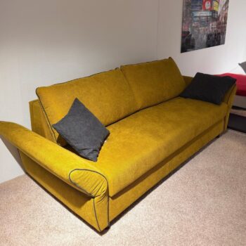 Ein senfgelbes „Signet Dauerschläfer“-Sofa mit zwei schwarzen Zierkissen steht auf einem beigen Teppich in der Nähe einer weißen Wand, über der ein gerahmtes Foto mit einer Stadtansicht von Kyoto-Go hängt.