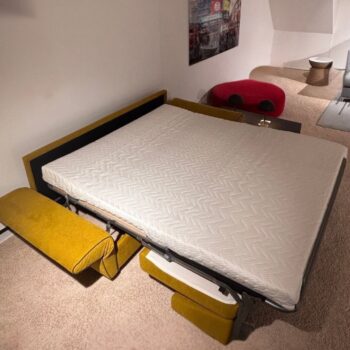 Ein gelbes „Dauerschläfer“-Schlafsofa ist ausgeklappt, die Matratze liegt in einem mit Teppichboden ausgelegten Raum frei. Im Hintergrund sind eine rote Ottomane und ein kleiner Beistelltisch zu sehen, die den vielseitigen Komfort der „Signet Kyoto-Go“-Kollektion unterstreichen.