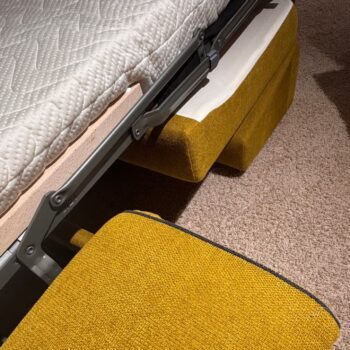 Eine Nahaufnahme eines gelb gepolsterten „Dauerschläfer“-Sofas von Signet, dessen Matratze teilweise ausgeklappt ist, sodass der robuste Metallrahmen und der strukturierte beige Teppich darunter zu sehen sind – ein einladendes Kyoto-Go-Design für jeden modernen Wohnraum.