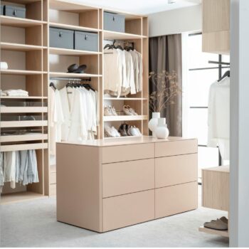 Begehbarer Kleiderschrank Fokus mit hellen Holzregalen, weißen Kleiderstangen und einer zentralen, hellrosa Inselkommode.