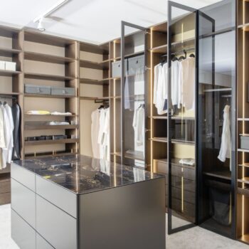 Ein begehbarer Kleiderschrank von Sudbrock mit Regalen aus dunklem Holz, Vitrinen mit Glasfronten und Innenbeleuchtung sowie einer zentralen Insel mit einer Arbeitsplatte aus dunklem Marmor.