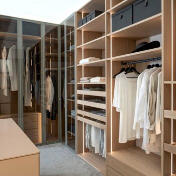 Ein begehbarer Kleiderschrank von Fokus mit hellen Holzregalen von Sudbrock, integrierter vertikaler LED-Beleuchtung, Schränken mit Glasfronten und hängender Kleidung.