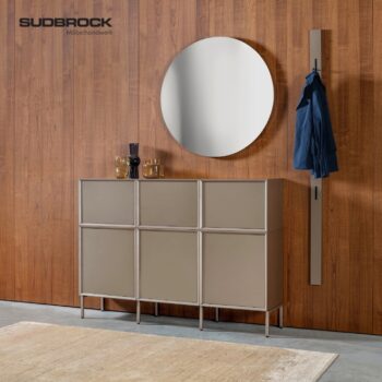 Ein moderner Eingangsbereich mit der eleganten Garderobe „Modo“ von Sudbrock, mit einem runden Spiegel, einem beigen Schrank, auf dem Gläser und Vasen stehen, einer blauen Jacke, die an einem Wandhaken hängt, und einer Holzwand, auf der das Logo von SUDBROCK Möbelhandwerk zu sehen ist.