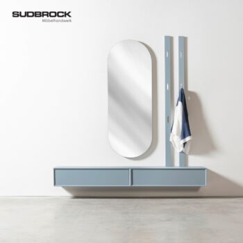 Eine moderne Wandkonsole aus der Modo Garderobe-Kollektion verfügt über zwei geräumige Schubladen, einen hohen ovalen Spiegel und zwei vertikale Kleiderhaken, an denen ein weiß-blaues Tuch hängt – ein perfektes Beispiel für Sudbrocks eleganten Ansatz beim funktionalen Garderobendesign.