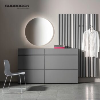 Minimalistisch eingerichteter Raum mit einem grauen Modo Garderobe-Kleiderschrank mit Schubladen, einem runden Spiegel, einer grauen Wandverkleidung aus Lamellen mit Haken, einem aufgehängten weißen Hemd, einem grauen Stuhl und zwei Büchern. Sudbrock-Logo oben links.