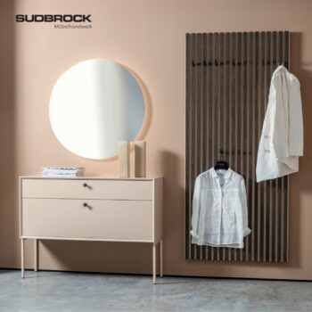 Minimalistischer Eingangsbereich mit einem runden Spiegel, zwei Büchern und einer beigen Modo Garderobe-Konsole mit Schubladen. Eine Wandverkleidung aus Latten dient als elegante Garderobe, an der zwei weiße Hemden hängen. An der Wand ist das SUDBROCK-Logo zu sehen.