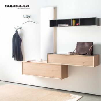 Moderner Eingangsbereich mit minimalistischen Wandschränken aus Holz aus der Kollektion „Sudbrock Modo Garderobe“, Kleiderhaken, an denen eine Jacke hängt, ein Regal mit Büchern, ein Spiegel und eine Ledertasche auf dem Schrank. Sudbrock Möbelhandwerk-Logo in der Ecke.