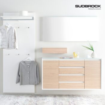 Moderner Eingangsbereich mit einem Garderobenschrank aus hellem Holz, einem Wandspiegel, Kleiderhaken und zwei aufgehängten Jacken; minimalistische Einrichtung mit einer grünen Pflanze und dem Logo von Sudbrock Möbelhandwerk in der Ecke.