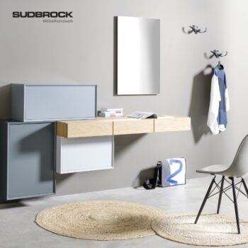 Ein moderner Eingangsbereich mit Wandschränken, einem schwebenden Schreibtisch, einem runden geflochtenen Teppich, einem Stuhl, einem Spiegel, Kleiderhaken und einem an der Wand hängenden weißen Hemd – alles in einer neutralen Farbpalette – schafft eine einladende Atmosphäre, die vom „Modo Garderobe“-Design von Sudbrock inspiriert ist.