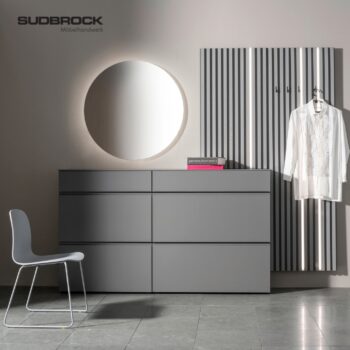 Modernes, minimalistisches Zimmer mit einer grauen Kommode, einem grauen Stuhl, einem runden Spiegel und einer Garderobenwand mit Haken, an denen ein weißes Hemd hängt; SUDBROCK Möbelhandwerk-Logo an der Wand.