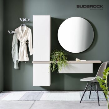 Minimalistischer Eingangsbereich mit weißen Hängeschränken, einem runden Spiegel, Garderobenhaken an der Wand, an denen ein Bademantel hängt, einem Stuhl und einer Topfpflanze vor einer grünen Wand. Ein Teppich und ein aufgeschlagenes Buch sind zu sehen. An der Wand ist die Marke SUDSBROCK neben Tando-Akzenten zu sehen.