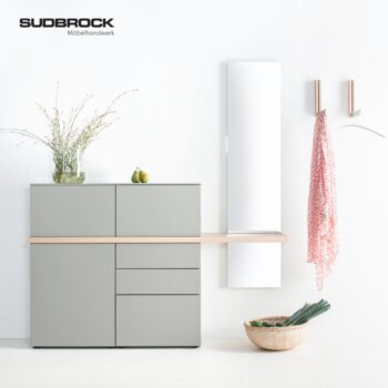 Minimalistischer Flur mit einem grauen Sudbrock-Garderobenschrank, einer dekorativen Vase, Äpfeln, einem Spiegel, Wandhaken, an denen ein roter Schal hängt, und einem Korb mit einem Lauch auf dem Boden. Sudbrock Möbelhandwerk-Logo oben links.