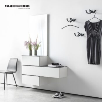 An einer Wand neben einem schwarzen Stuhl sind weiße, moderne, freitragende Schubladen von Garderobe, ein Spiegel und eine Vase mit Blumen angebracht. An Wandhaken hängt ein schwarzes Kleid. Auf dem Boden stehen weiße Sneaker, die dieses elegante, von Udbrock inspirierte Arrangement abrunden.