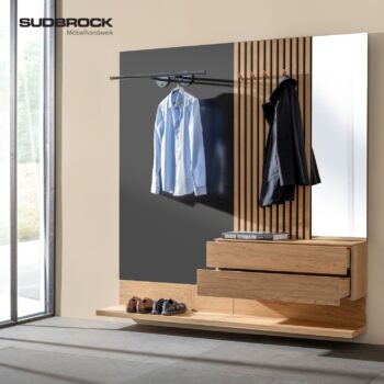 Moderne Garderobe für den Eingangsbereich mit Holzschubladen, einem Schuhregal, Kleiderhaken, einer Kleiderstange für Hemden und einem großen Spiegel. In der oberen linken Ecke ist das Logo von Subbock Möbelhandwerk zu sehen. Inspiriert von den „Tando“-Designelementen von udbrock vereint dieses Möbelstück Stil mit praktischen Aufbewahrungslösungen.