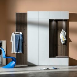 Moderne Eingangsgarnitur mit weißen Schränken, vertikalen Holzpaneelen und praktischen Garderobenelementen, bestehend aus zwei aufgehängten Hemden, einem blauen Stuhl, einem Sitzkissen und einer aufgeschlagenen Zeitschrift auf dem Boden. Vor einem neutralen Wandhintergrund sorgt sie für einen klaren, einladenden Look.