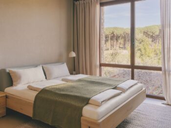 Minimalistisches Schlafzimmer mit einem Holzbett, einer grünen Decke und einem großen Fenster mit Blick auf Bäume. Beigefarbene Vorhänge und von Nox inspirierte neutrale Farben an Wänden und Dekoration schaffen einen friedlichen, beruhigenden Rückzugsort.