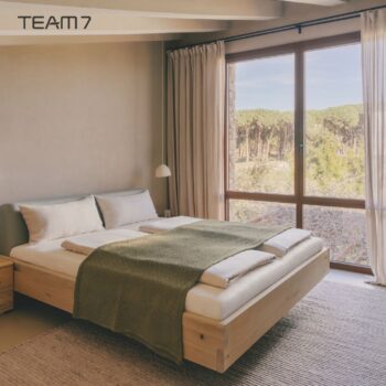 Minimalistisches Schlafzimmer mit einem Holzbett, einer grünen Decke und einem großen Fenster mit Blick auf Bäume. Beigefarbene Vorhänge und von Nox inspirierte neutrale Farben an Wänden und Dekoration schaffen einen friedlichen, beruhigenden Rückzugsort.