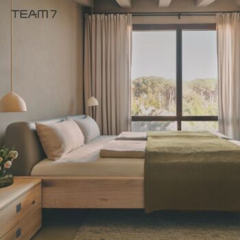 Minimalistisches Schlafzimmer mit einem Nox-Holzbett, heller Bettwäsche, zwei Hängelampen, einem Nachttisch mit Blumen und einem großen Fenster mit Blick auf Bäume. Das TEAM 7-Logo befindet sich in der oberen linken Ecke.