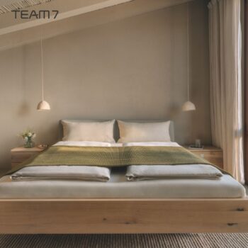 Ein ordentlich gemachtes Bett mit hellgrauer NOX-Bettwäsche und einer grünen Decke, flankiert von zwei hölzernen Nachttischen und hängenden Pendelleuchten in einem modernen, minimalistischen Schlafzimmer.