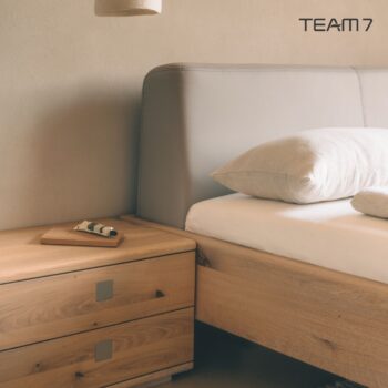 Minimalistische Schlafzimmerszene mit einem hölzernen Nox-Nachttisch, einem Bett mit beigem Kopfteil und Bettzeug sowie einem kleinen Gegenstand auf dem Nachttisch. Das TEAM 7-Logo ist in der oberen rechten Ecke zu sehen.