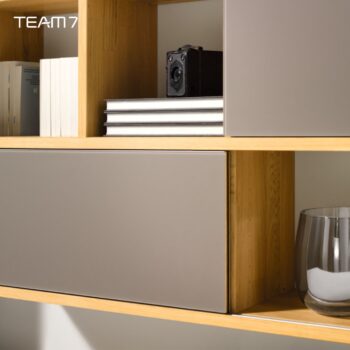 Ein hölzernes Cubus-Regal fasst Bücher, einen Stapel von vier Notizbüchern, eine alte Kamera und eine Metalltasse. Das Wort TEAM 7 erscheint in der linken oberen Ecke und spiegelt die Eleganz und das funktionale Design von Wohnwand wider.