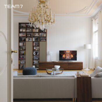 Ein modernes Wohnzimmer mit einem grauen Sofa, einem Cubus-Holzregal, einem Fernseher mit Kunstwerken, einem Kronleuchter, einer Stehlampe und großen Fenstern, die natürliches Licht hereinlassen. TEAM 7-Logo in der oberen linken Ecke.