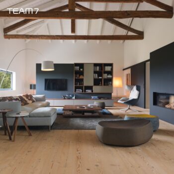 Modernes Wohnzimmer mit sichtbaren Holzbalken, neutralen Möbeln, großen Fenstern und einem gemütlichen Kamin. Eine elegante Team 7 Wohnwand aus der Cubus-Kollektion bietet eingebaute Regale, die den hellen Holzboden und die minimalistische Einrichtung ergänzen.