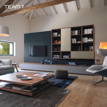 Modernes Wohnzimmer mit Holzböden, einer Team 7 Wohnwand aus der Cubus-Linie, wandmontiertem TV, Regalen mit Büchern und Dekoration, einem Couchtisch und modernen Sitzgelegenheiten unter freigelegten Deckenbalken.
