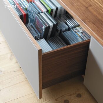 Eine offene Holzschublade in einem modernen Cubus-Schrank von Team 7 zeigt CDs und DVDs übersichtlich nach ihren Rücken geordnet auf einem hellen Holzboden - die perfekte Ergänzung zu Ihrer Wohnwand.