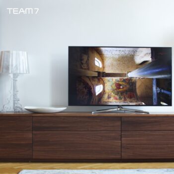 Ein Flachbildfernseher auf einem Cubus-Holzschrank von Team 7 zeigt ein Bild einer bemalten Decke und Säule; eine klare Lampe und eine dekorative Schale sitzen neben dem Fernseher und werten diese moderne Wohnwand auf.