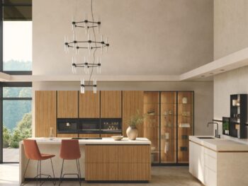 Moderne Küche mit Team7-Küchenschränken aus Holz, einer zentralen Insel mit zwei orangefarbenen Stühlen, einem minimalistischen Kronleuchter, großen Fenstern und neutral getönten Wänden für eine einladende, moderne Atmosphäre.