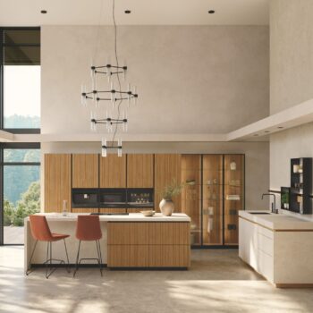 Moderne Küche mit Team7-Küchenschränken aus Holz, einer zentralen Insel mit zwei orangefarbenen Stühlen, einem minimalistischen Kronleuchter, großen Fenstern und neutral getönten Wänden für eine einladende, moderne Atmosphäre.