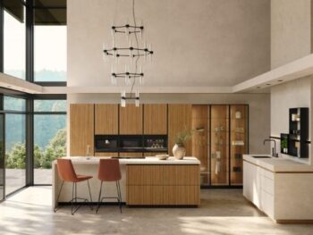 Moderne Küche mit großen Fenstern, Holzschränken, einer zentralen Insel mit zwei Stühlen, Einbaugeräten und minimalistischen Pendelleuchten. Der Außenbereich ist durch die Glaswände sichtbar.