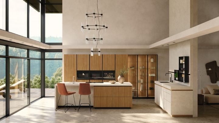 Moderne Küche mit großen Fenstern, Holzschränken, einer zentralen Insel mit zwei Stühlen, Einbaugeräten und minimalistischen Pendelleuchten. Der Außenbereich ist durch die Glaswände sichtbar.