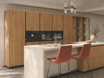 Moderne Team7 Küche mit Holzschränken, einer hellen Steininsel, zwei orangefarbenen Barhockern, Gläsern und einer dekorativen Vase unter einem schwarz-silbernen Kronleuchter - verleihen Sie Ihrem Raum den eleganten echt.zeit evo touch.
