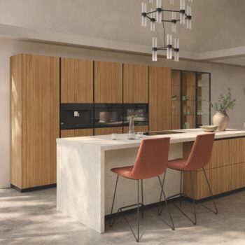 Moderne Team7 Küche mit Holzschränken, einer hellen Steininsel, zwei orangefarbenen Barhockern, Gläsern und einer dekorativen Vase unter einem schwarz-silbernen Kronleuchter - verleihen Sie Ihrem Raum den eleganten echt.zeit evo touch.