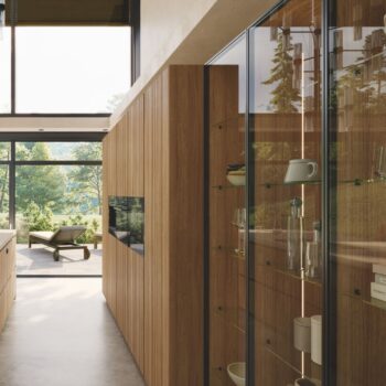 Modernes Interieur mit Holzvertäfelung, Glasregalen und großen Fenstern mit Blick auf einen grünen Außenbereich mit Lounge-Sesseln - mit eleganten Team7-Küchenelementen für einen raffinierten Touch.
