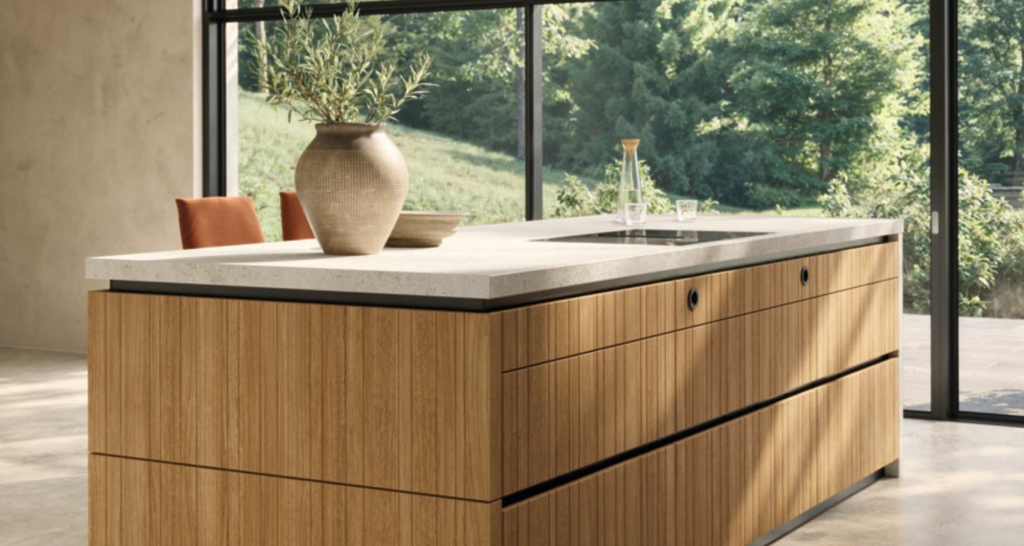 Eine moderne Team7-Kücheninsel mit Holzverkleidung und einer hellen evo-Arbeitsplatte steht in einem großzügigen Raum mit großen Fenstern und Blick auf eine grüne Außenlandschaft - perfekt, um Momente der echt.zeit zu schaffen.
